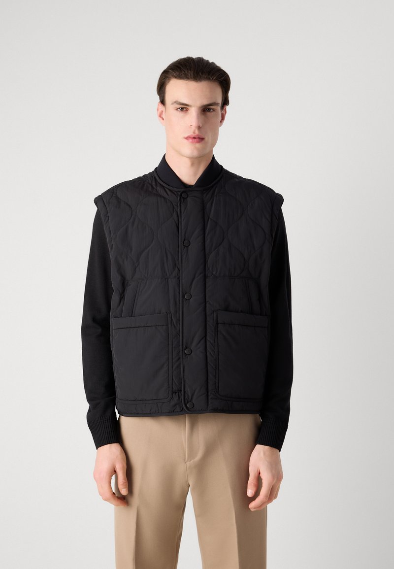 Schwarze gesteppte Weste mit hohem Kragen, vorderen Druckknöpfen und zwei großen Taschen. Getragen über einem schwarzen Langarmshirt und beigen Hosen.
