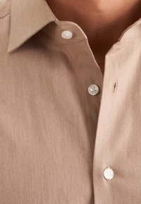 Camicia marrone con bottoni, realizzata in tessuto testurizzato, con colletto a punta e bottoni bianchi, con una piccola lacerazione all'apertura.