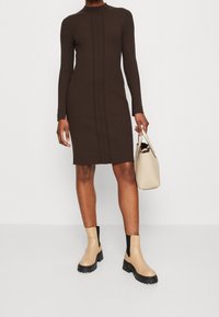 Femme portant une robe marron côtelée à manches longues, tenant un sac à main beige, et chaussant des bottines beige avec des panneaux élastiques noirs.