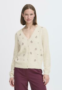 Cardigan color crema con scollo a V, chiusura con bottoni e ricamo floreale. Tessuto morbido con maniche lunghe e orlo a coste. Abbinato a pantaloni scuri.