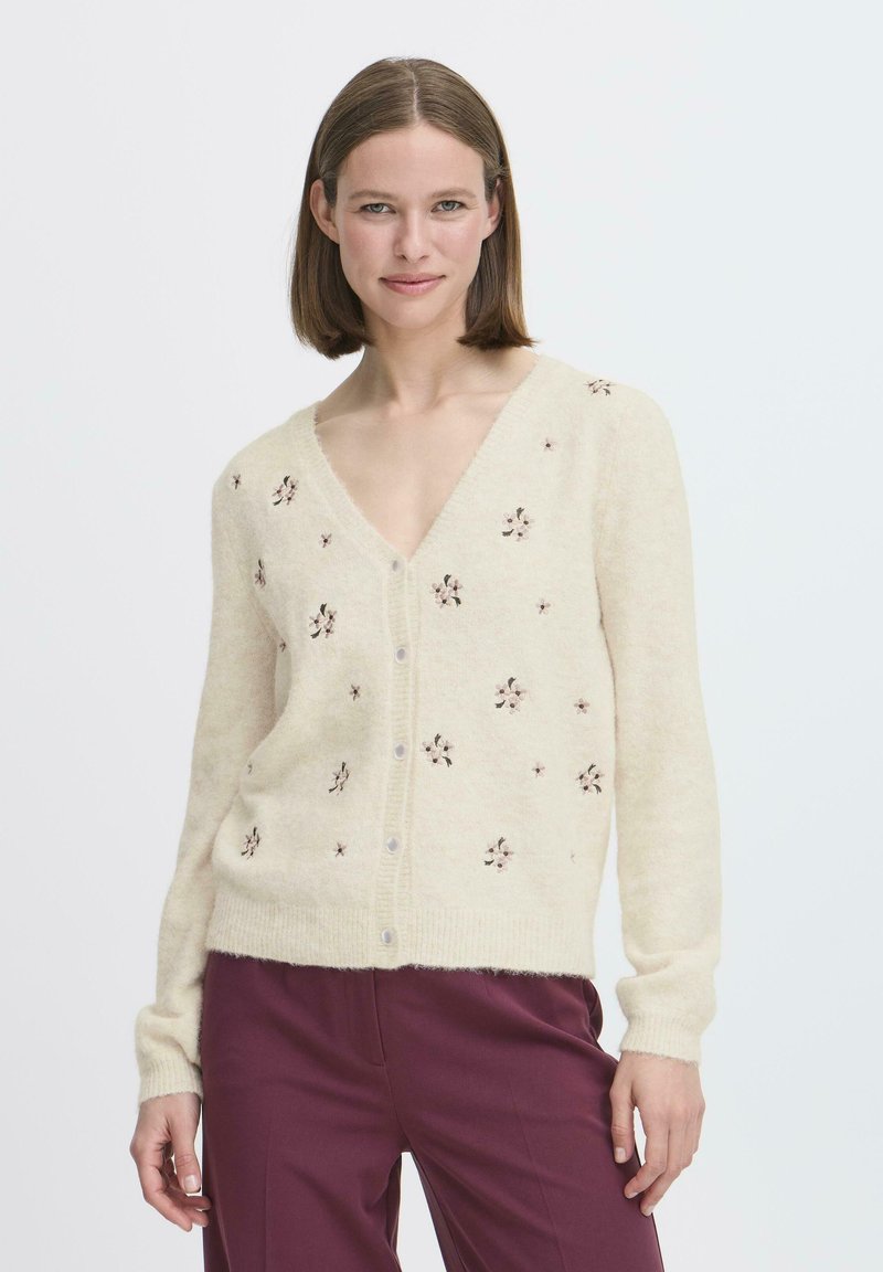 Cardigan color crema con scollo a V, chiusura con bottoni e ricamo floreale. Tessuto morbido con maniche lunghe e orlo a coste. Abbinato a pantaloni scuri.