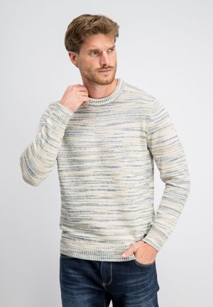 Man met lichtbruin haar, gekleed in een crème en blauw gestreepte gebreide trui en donkerblauwe jeans, die zijn nek aanraakt en opzij kijkt.