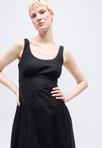 Γυναίκα με κοντά ξανθά μαλλιά, ντυμένη με μαύρο φόρεμα χωρίς μανίκια, στέκεται με τα χέρια στη μέση μπροστά σε απλό φωτεινό φόντο.