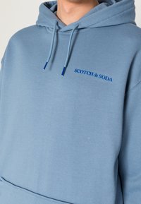 Sudadera azul claro con bolsillo tipo canguro, capucha con cordones y el logo de SCOTCH & SODA bordado en azul en contraste en el pecho.