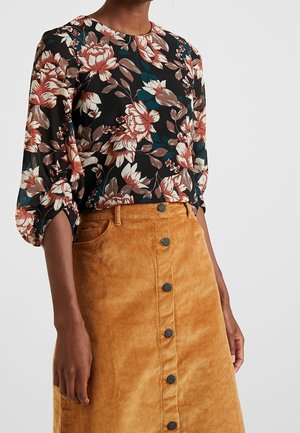 Kvinde iført en sort blomsterbluse med pufærmer og et højtaljet, lysebrunt fløjlsnederdel med knapper foran.