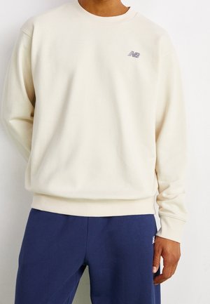 Mężczyzna w kremowej bluzy z okrągłym dekoltem z małym logo New Balance oraz granatowych spodniach dresowych na jednolitym tle.