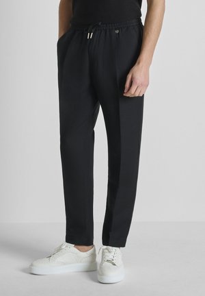 Personne portant un pantalon noir à cordon et des baskets blanches à lacets, debout avec une main dans la poche sur un fond gris uni.