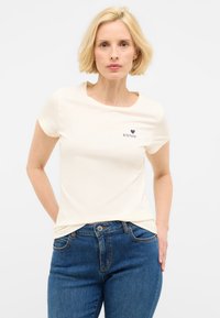 Camiseta de manga corta color crema con escote redondo y pequeño logo azul "MUSTANG" en el pecho, combinada con jeans de mezclilla azules.