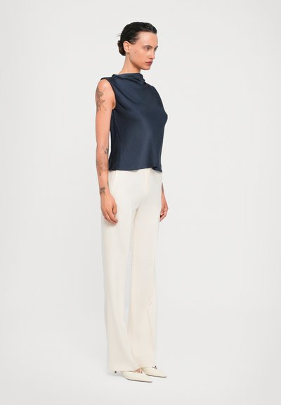 Navyfarbener ärmelloser Top aus glattem Stoff, kombiniert mit weißen, weit geschnittenen Hosen. Schlanker Schnitt mit hohem Hals und minimalistischen Design.