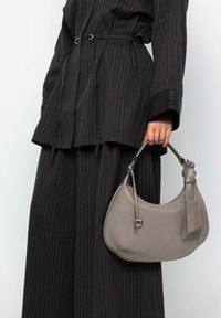 Sac à main en cuir gris avec une forme incurvée, un design léger et une anse nouée, tenu par une personne vêtue d'une tenue noire à rayures.