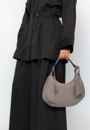 Graue Lederhandtasche mit geschwungener Form, leichter Bauweise und geknotetem Griff, gehalten von einer Person in einem schwarz gestreiften Outfit.