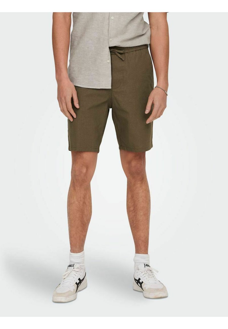 Olivgrüne Shorts aus Baumwolle mit lockerer Passform, Kordelzugbund und mittellanger Länge bis zum Oberschenkel. Tragen mit weißen Sneakers.