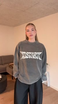 Šedá oversized mikina s bílým grafickým nápisem, dlouhými rukávy a kulatým výstřihem, kombinovaná s černými kalhotami s širokými nohavicemi. Ležérní materiál a střih.