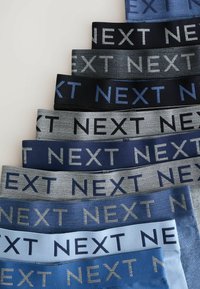 Egy csomag rugalmas derekú övekből, kék, szürke és fekete különböző árnyalataiban, a "NEXT" szóval kontrasztos betűkkel.
