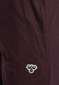Bordowe spodnie z materiału z boczną kieszenią i haftowanym logo w czerni i bieli. Gładka tekstura z widocznymi detalami ściegów.