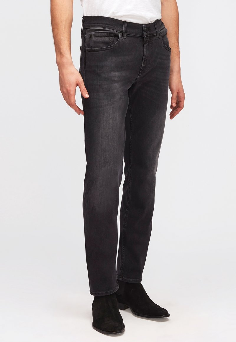 Homme portant un jean noir slim-fit et des bottines noires en daim, montré de la taille aux pieds sur fond blanc.