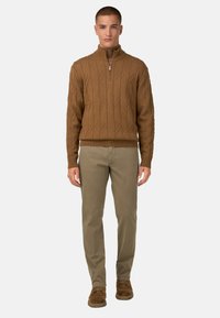 Brauner Kabelstrickpullover mit einem Halbreissverschluss-Kragen, kombiniert mit olivgrünen Hosen und braunen Wildlederschuhen. Neutrale Farbpalette und strukturiertes Design.