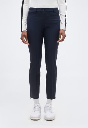 Model feminin purtând pantaloni albastru închis, mulați pe gleznă, șosete albe, pantofi sport albi și un top alb cu mâneci lungi și dungi negre pe mâneci.