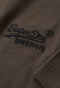 Tejido marrón con una textura suave que presenta el logotipo bordado en negro "SuperDry" y una línea de acento curva, cosida en el lado izquierdo.