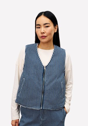 Blauw-wit gestreept vest met een ritssluiting aan de voorkant, gemaakt van stof die lijkt op denim, met brede armsgaten en zijzakken. Draagt over een wit shirt.