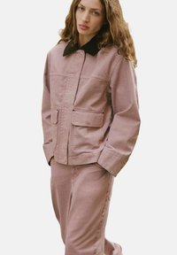 Giacca in tela rosa con colletto nero, dotata di due tasche frontali e chiusura con zip, abbinata a pantaloni rosa coordinati.