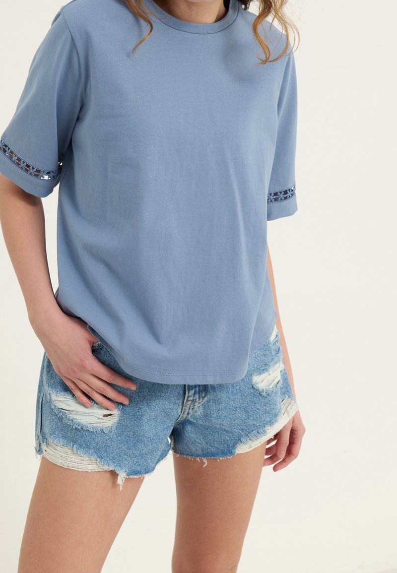 Femme portant une chemise à manches courtes bleu clair avec bordure en dentelle et un short en jean déchiré sur un fond uni.