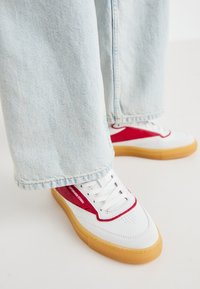 Hvite sneakers med røde detaljer, med hull i tåboksen, hvite lisser og en gummisåle. Brukt med lyseblå løstsittende jeans.