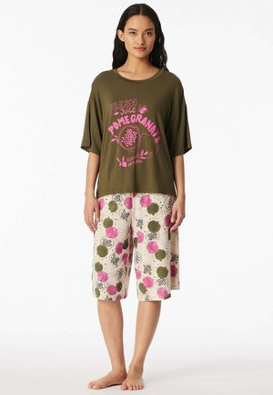 Woman wearing olive green loose t-shirt with pink "Pomegranate" print and knee-length cream shorts with pink and green floral patterns.