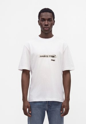 Jeune homme portant un t-shirt blanc avec le texte "Favourite T-Shirt Hugo" et un jean bleu, debout devant un fond blanc uni.