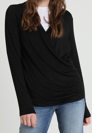 Kobieta ubrana w czarny, długi rękaw typu wrap na białej koszuli, w połączeniu z jasnoniebieskimi dżinsami, widoczna od szyi do bioder.