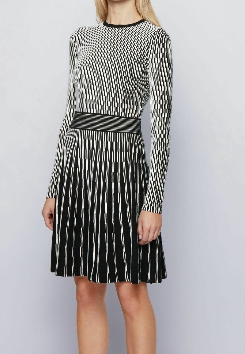 Femme portant une robe noire et blanche à manches longues avec motif géométrique et ceinture rayée, debout devant un fond clair et uni.