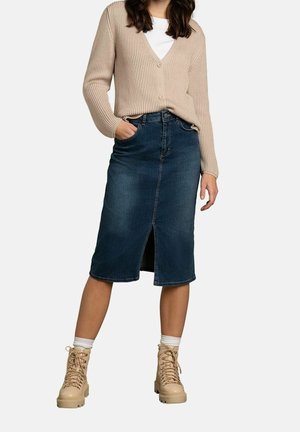 Kvinde iført beige cardigan, hvid top, mørkeblå jeansnederdel til knæet med frontslids, hvide sokker og beige snørestøvler.