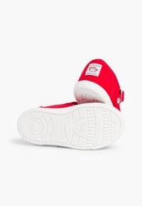 Zapatillas de lona rojas con suelas de goma blanca y hebilla lateral. Presenta un parche con un logo de mono en el talón. Textura suave y diseño sencillo.