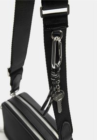 Tracolla in tessuto nero con hardware argentato, caratterizzata da un dettaglio chiave e finiture con cerniera sulla borsa nera attaccata.