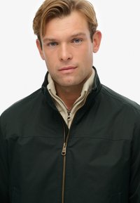 Chaqueta verde oscura con cuello alzado, con un zipper dorado y forro beige, textura lisa y un diseño sencillo.