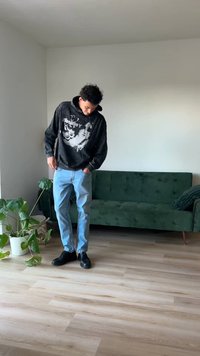 Hanorac negru cu imprimeu grafic, blugi de culoare albastru deschis și încălțăminte neagră. Canapea din catifea verde și plantă în ghiveci în fundal. Podea din lemn deschis.