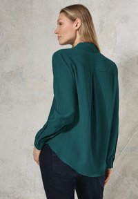 Blusa texturizzata verde acqua con vestibilità morbida, maniche lunghe e polsini a bottone. La parte posteriore presenta una cucitura a yoke e un orlo leggermente curvo.