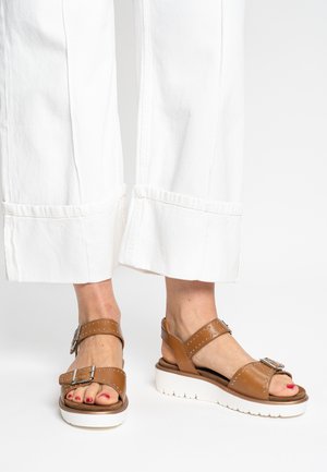 Brown leather sandals with two adjustable straps, silver studs, and a white, textured platform sole. Paired with white wide-leg pants.