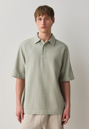Ung mand iført en lys grøn tekstureret kortærmet polo-shirt og beige teksturerede bukser, der står mod en ensfarvet baggrund.