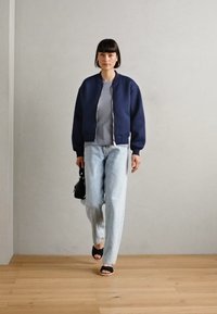 Marine bomberjakke, grå topp, lyseblå jeans, svarte sandaler. Modellen holder en liten svart håndveske, går på et tregulv mot en grå vegg.