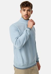 Lyseblå pullover med høj krave, med lange ærmer og ribbede manchetter. Accentueret med mørke striber på kraven og kanten.