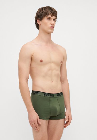 Zeleni pamučni bokserice s crnim "MOSCHINO" pojasom, koji imaju prilagodljiv oblik i glatku teksturu. Model je pozirao bočno uz bijelu pozadinu.