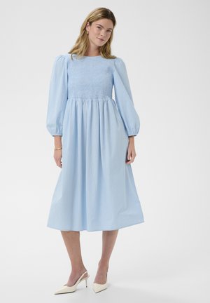 Femme portant une robe midi bleu clair avec des manches longues bouffantes et un corsage froncé, associée à des escarpins beige à bride arrière et des boucles d'oreilles créoles dorées.