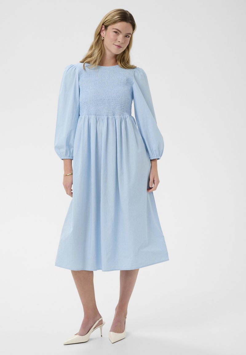 Frau trägt ein hellblaues Midikleid mit gepufften langen Ärmeln und gerafftem Oberteil, kombiniert mit beigen Slingback-Pumps und goldenen Creolen.