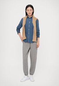 Modrá rifľová košeľa s gombíkmi, doplnená béžovou pletenou vestou. Šedé tepláky a biele tenisky dokončujú outfit.