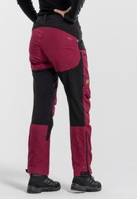 Waterproof outdoor pants in burgundy and black. Features reinforced areas, multiple pockets, and adjustable cuffs. Material appears durable.