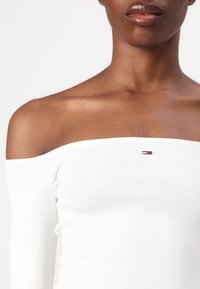 Bela bluzica z off-shoulder rezom z dolgimi rokavi, ribana tekstura, z majhnim rdečim, belim in modrim logotipom na prsih.