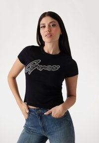 Černé žebrované tričko s krátkým rukávem, které má stříbrné kovové logo "Guess" umístěné na hrudi, stylizované s modrými džíny.