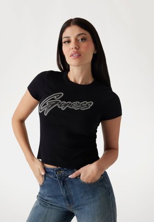 Zwarte ribbel T-shirt met korte mouwen, voorzien van een zilverkleurig metallic "Guess" logoontwerp op de borst, gecombineerd met blauwe spijkerbroek.