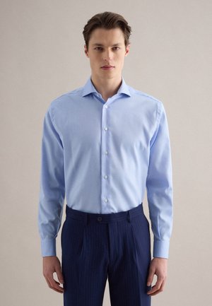 Uomo con una camicia azzurro chiaro abbottonata infilata in pantaloni blu scuro a righe sottili, in piedi davanti a uno sfondo beige uniforme.